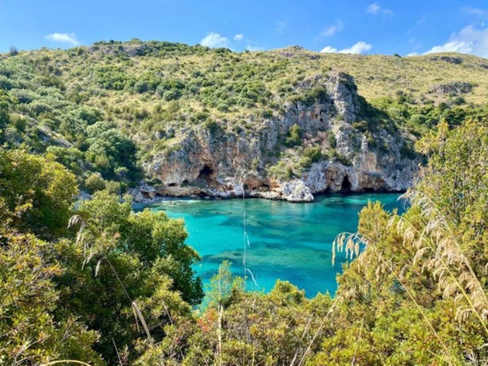 A clear turquoise bay surrounded by rocky cliffs and lush green vegetation under a blue sky with scattered clouds.