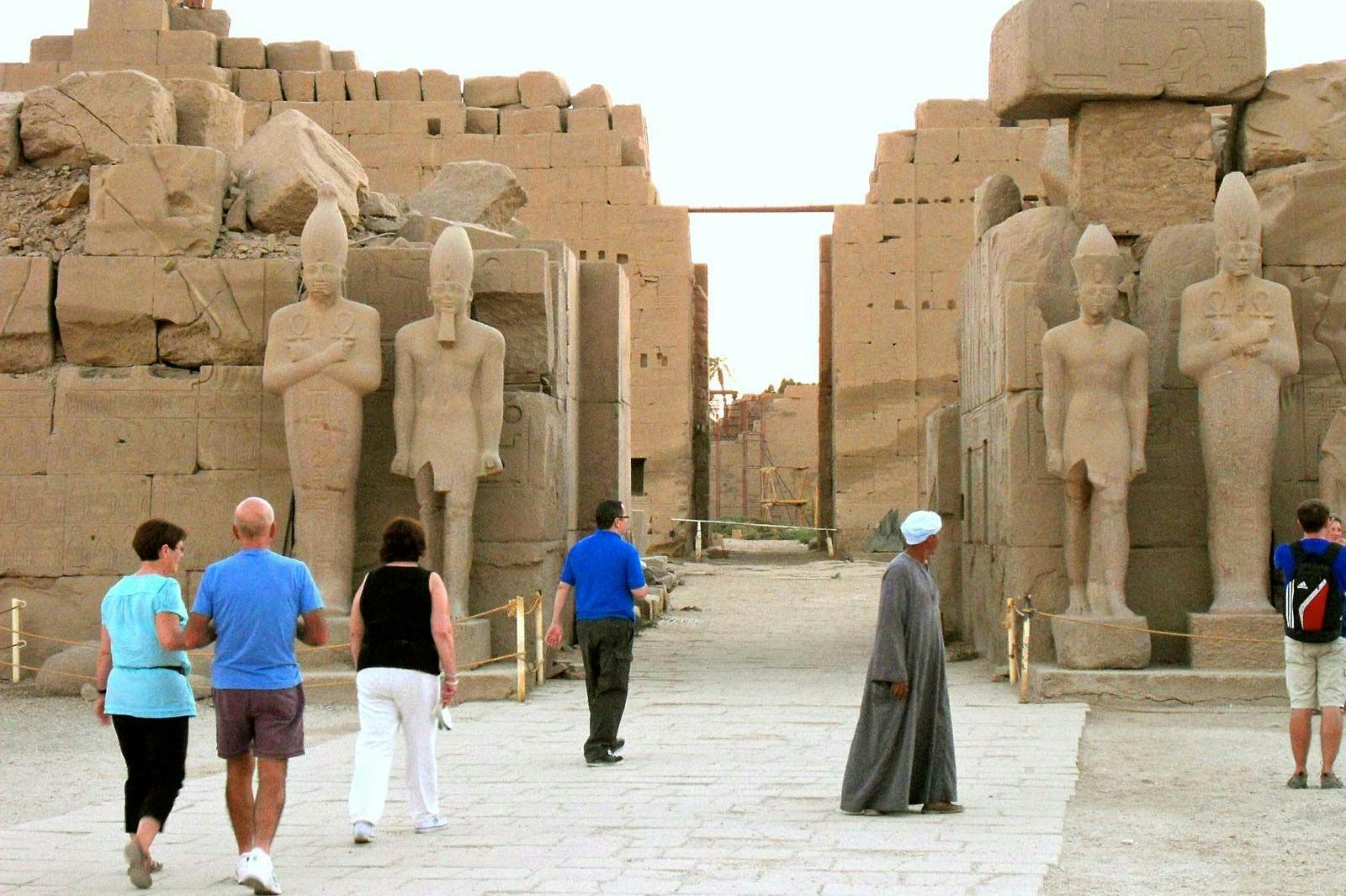 People walking through the ruins of a temple complex with large Egyptian statues and ancient stone structures.
