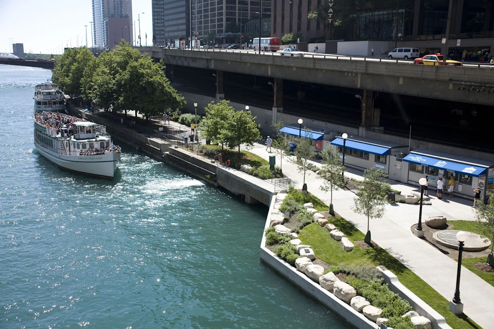 A riverside cityscape with a boat on the water, a path with trees and benches, and elevated roads and buildings in the background.