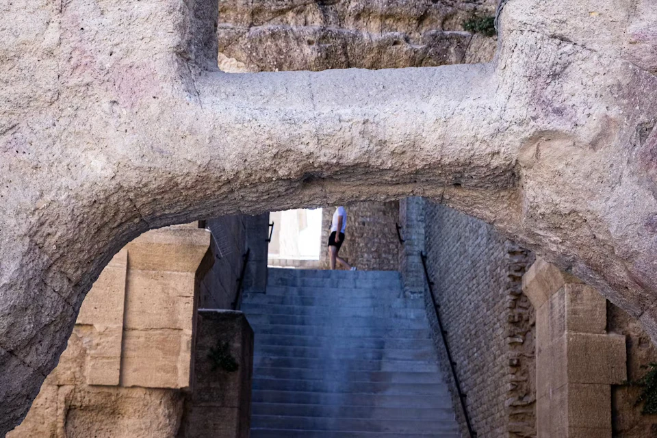 A person in shorts walks up a long stone staircase under a large stone archway, with ancient brick walls on both sides.