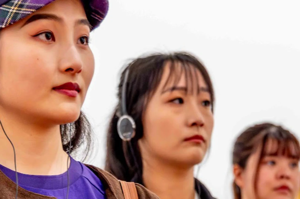 Three women wearing headphones stand in a bright room, likely observing something, with focused expressions.