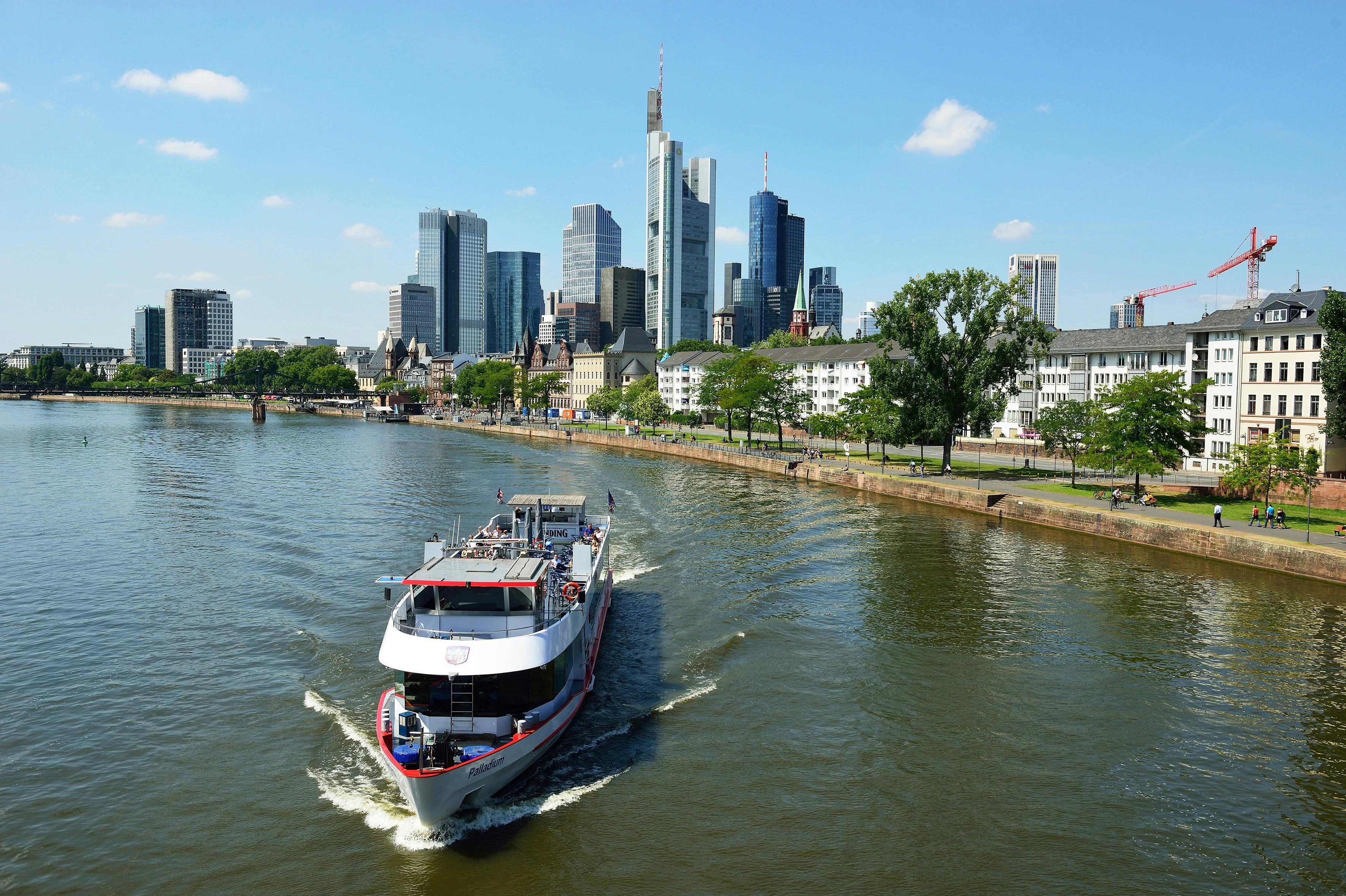 A boat sails on a river with a backdrop of city skyscrapers, older buildings, and a riverside park with people walking.