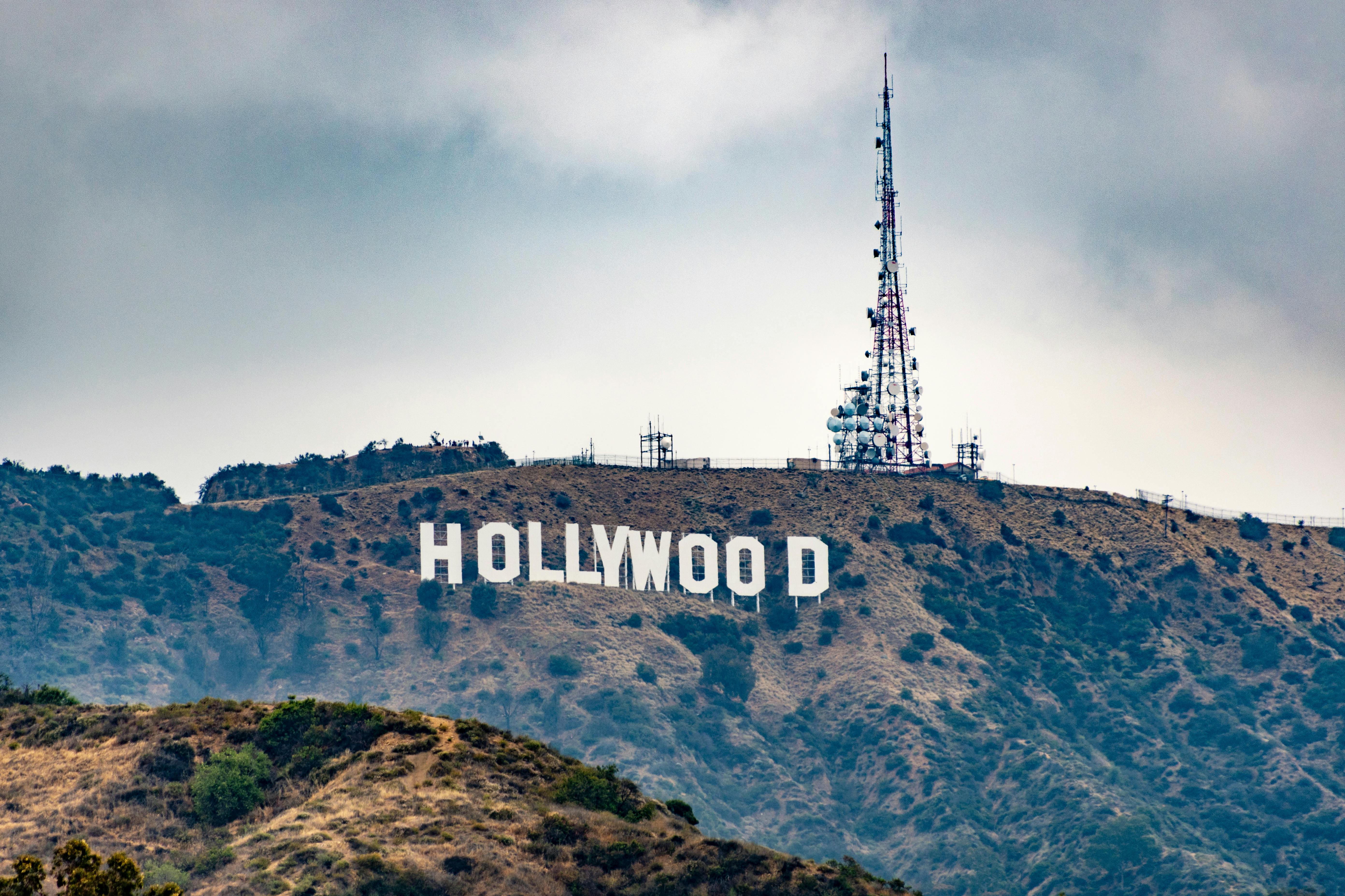 The Hollywood Sign on a hillside, with a transmission tower beside it against a cloudy sky.