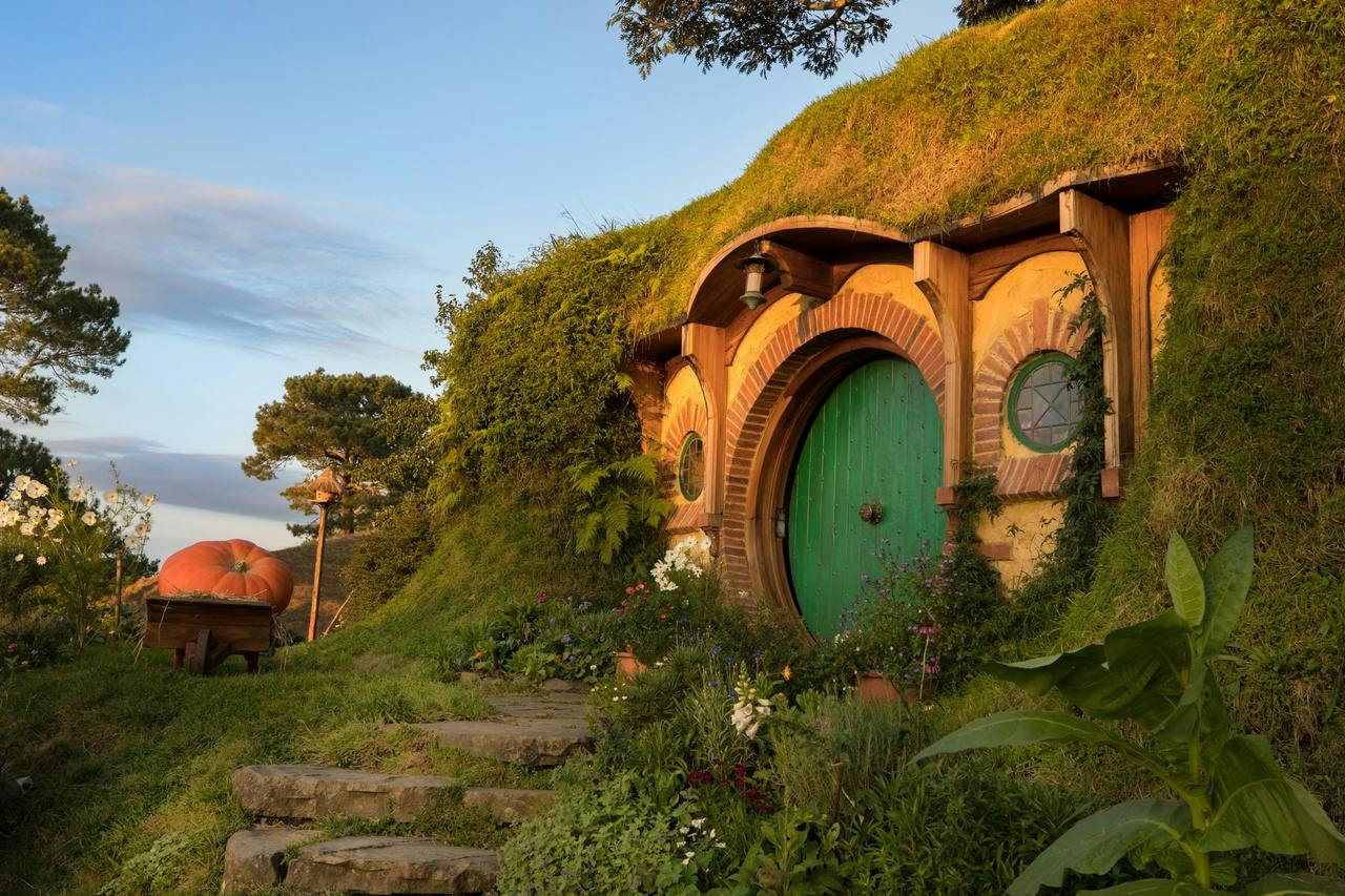 A small, round, green door built into a grassy hill, surrounded by plants and flowers in warm evening light.