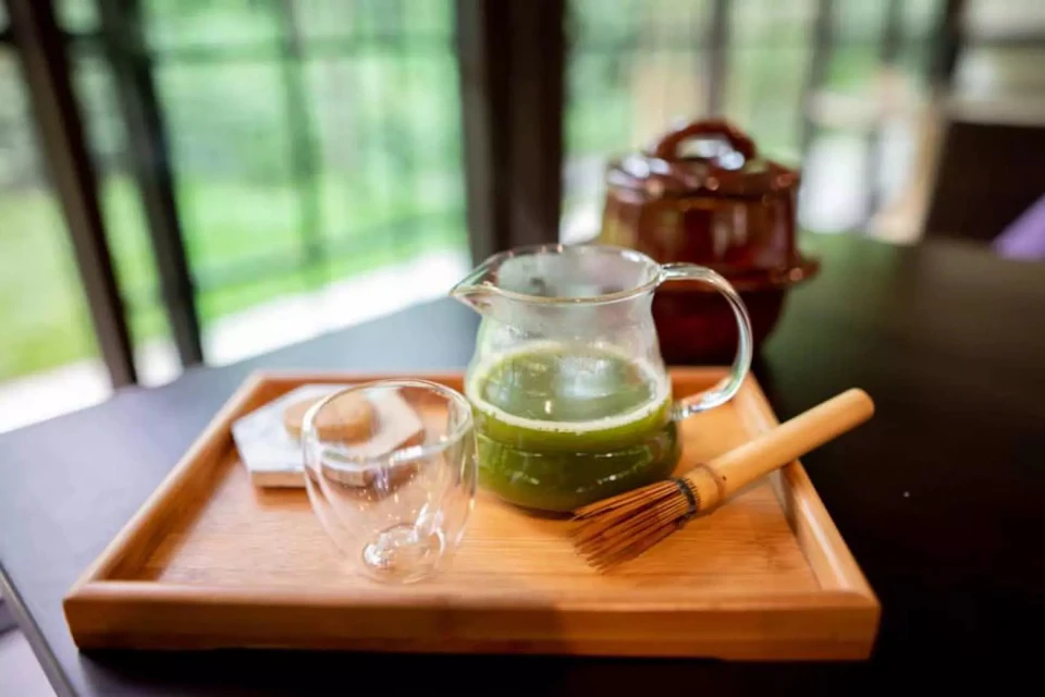 A tray with a glass pitcher of green tea, a whisk, a small glass cup, and a napkin on a table near a window.