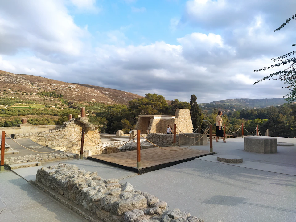 An ancient ruins site with stone structures, a wooden walkway, and two people walking against a backdrop of hills and cloudy sky.