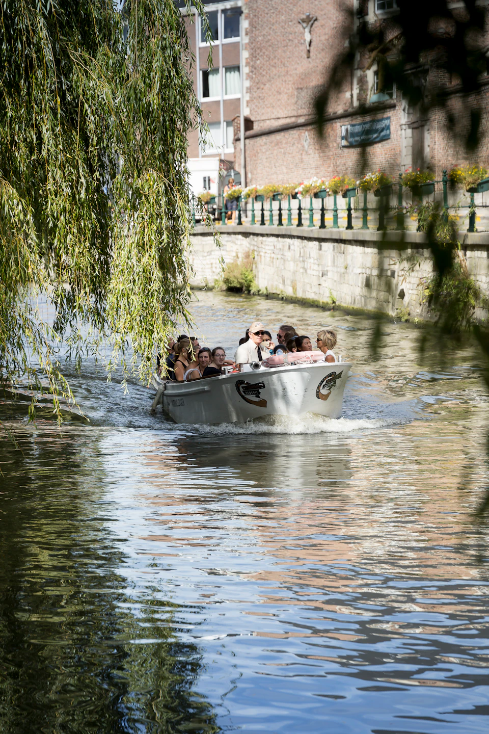 A group of people enjoy a boat ride on a calm canal, with weeping willow branches hanging down and a brick-lined shore nearby.