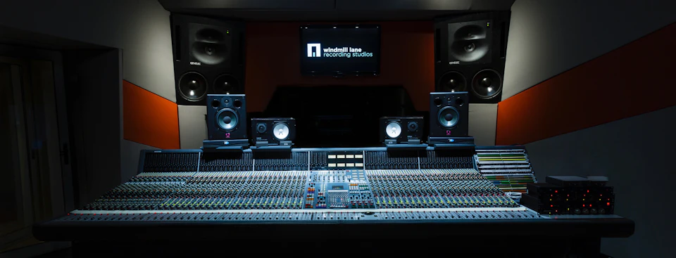 Close-up of a recording studio mixing console with multiple dials and switches, large speakers, and a screen displaying "Windmill Lane Recording Studios."
