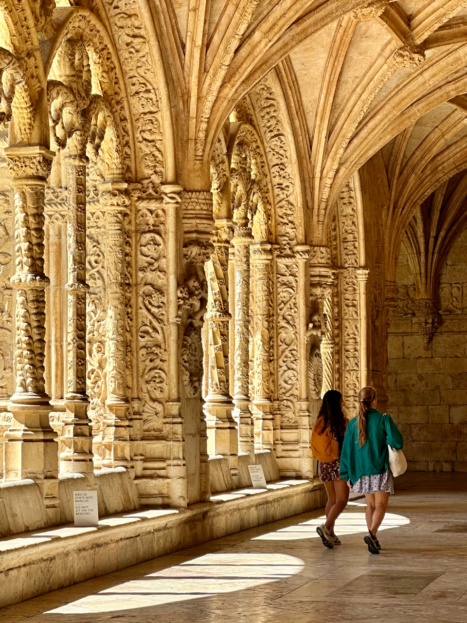 Two people walking through a stone corridor with ornate, carved arches and detailed columns, bathed in warm sunlight.