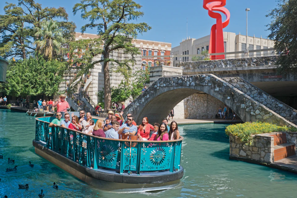 A group of people on a turquoise boat ride along a canal, with a stone bridge and a red sculpture in the background.