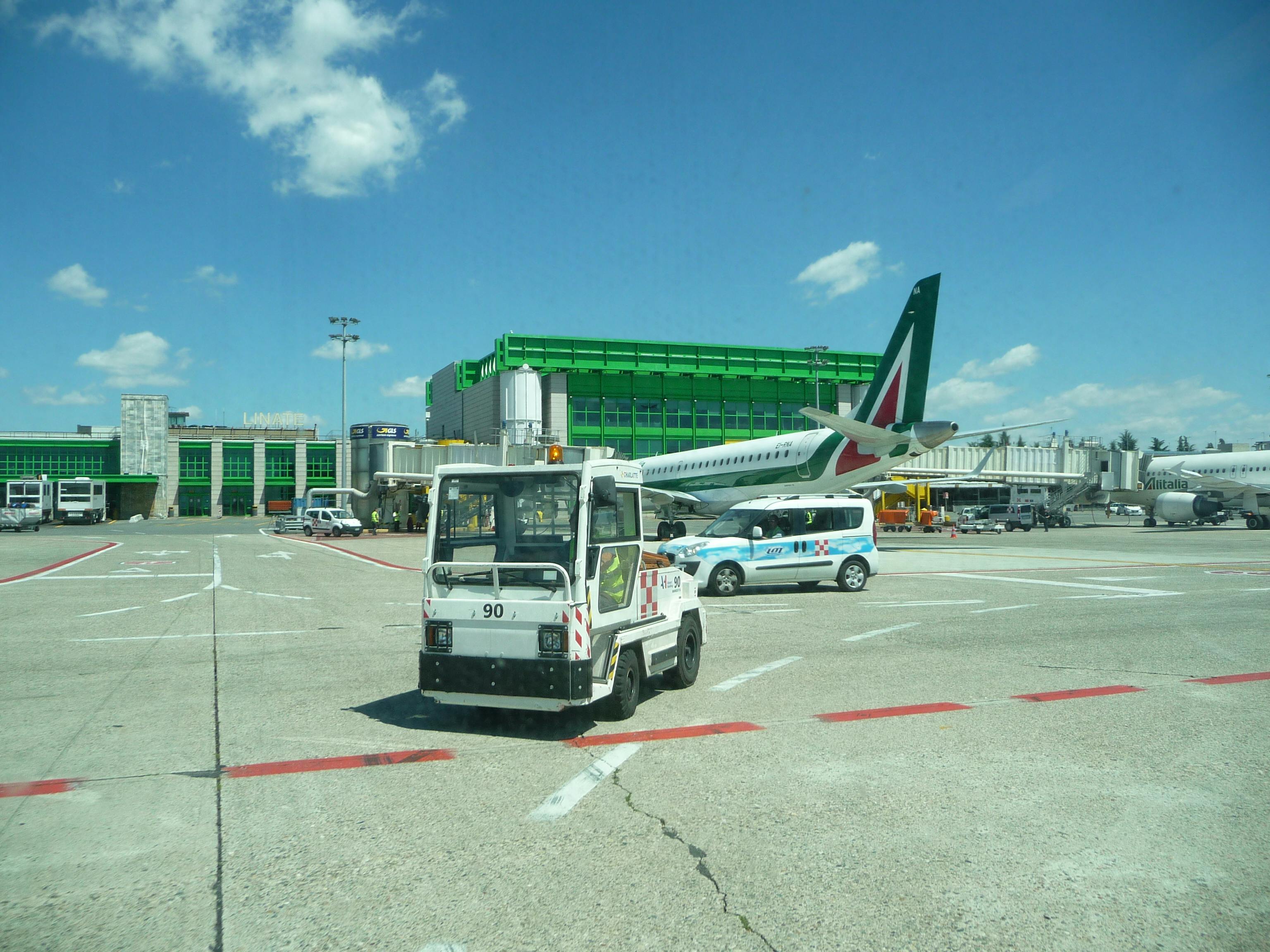 An airport tarmac with a small utility vehicle, a parked airplane, service vehicles, and a terminal building in the background.