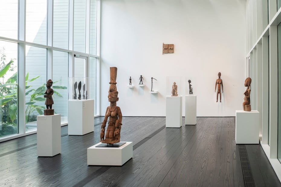 커다란 창문과 흰색 벽 근처에 있는 받침대와 선반에 아프리카 조각품이 전시된 미술관.