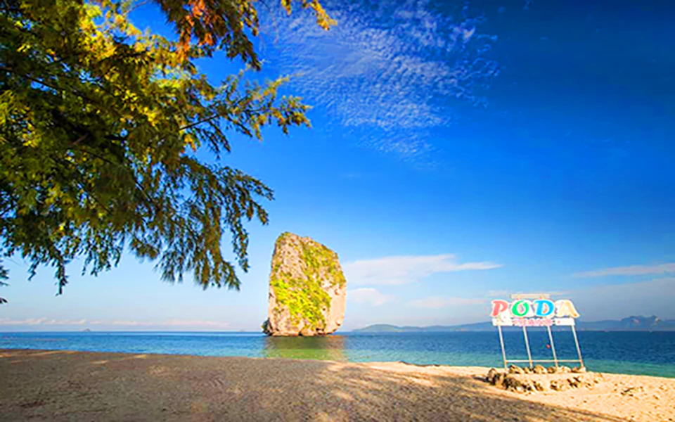 A sandy beach with a tree, clear blue sky, a rock formation in the sea, and a sign with partially visible text "PO..." on the right.