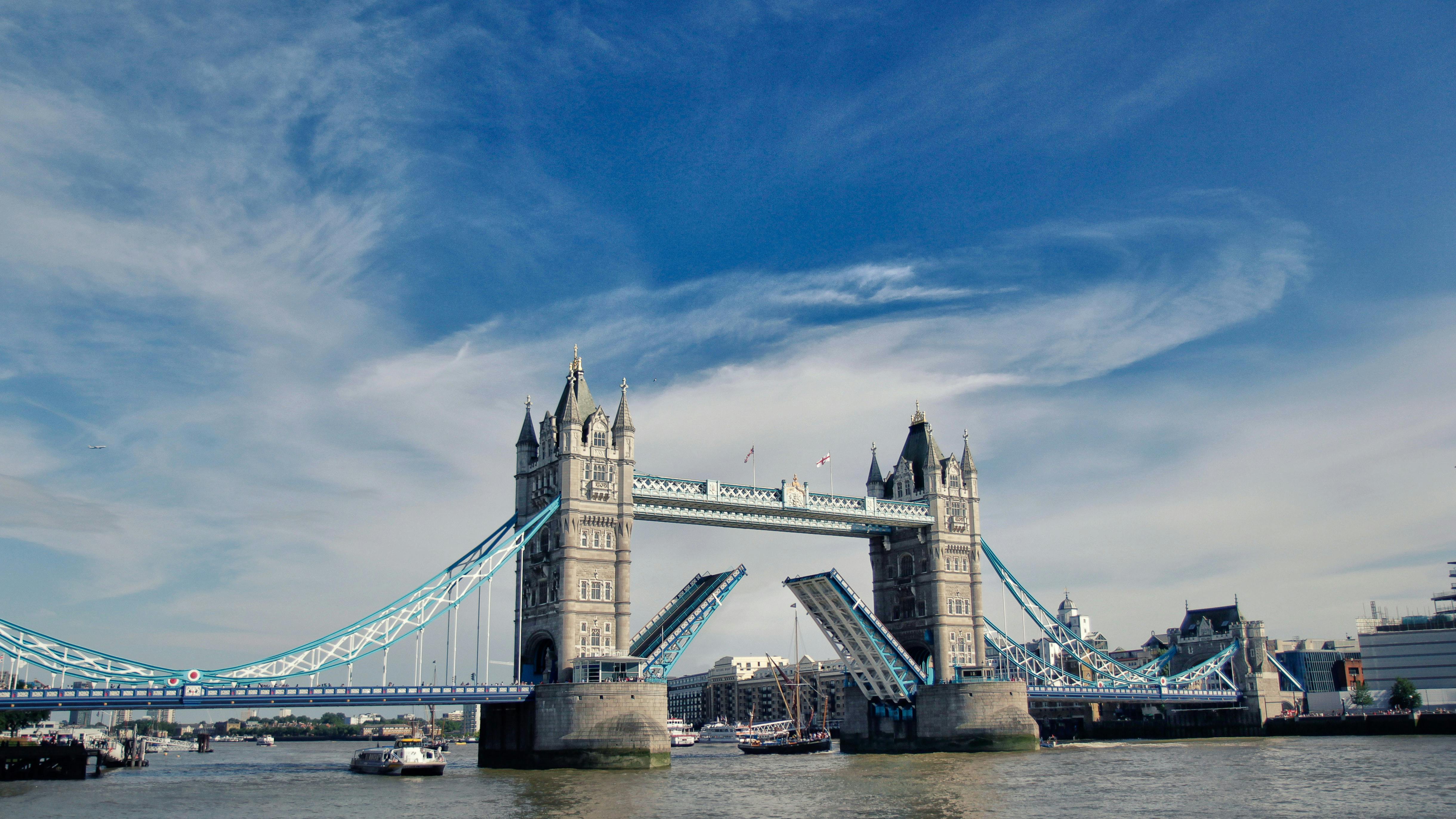 런던의 타워 브릿지 (Tower Bridge) 는 바스큘 (bascules) 이 높이 솟아 있고, 보트가 아래에서 항해하고 있으며, 머리 위로 구름이 흩어져 있는 밝고 푸른 하늘을 자랑합니다.