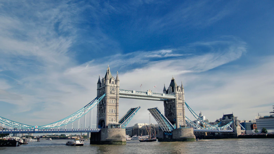 Tower Bridge in London with its bascules raised, boats sailing underneath, and a bright blue sky with scattered clouds overhead.