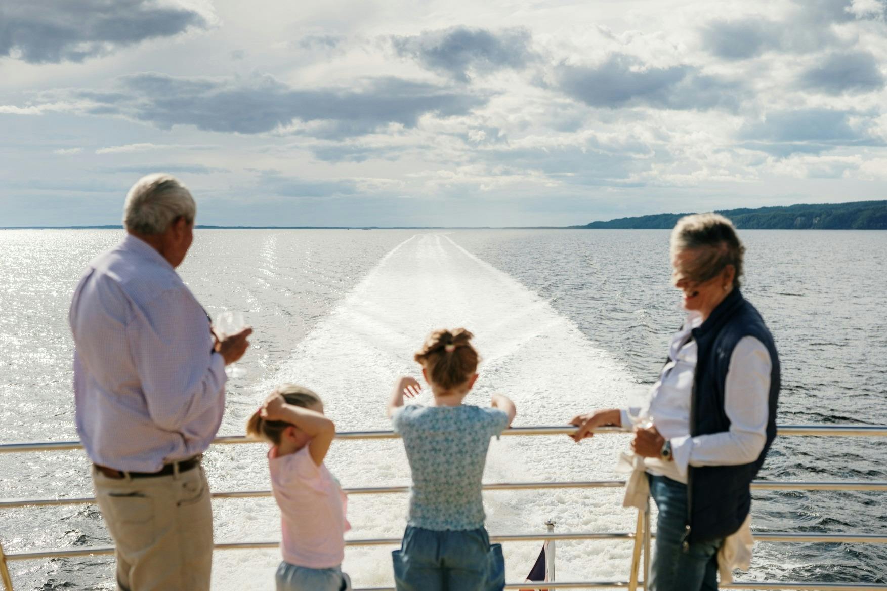 A group of people on a boat deck looking at the wake trailing behind, with water and distant land under a cloudy sky.
