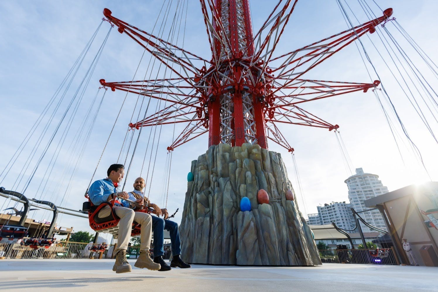 Two people seated on a swing ride, part of a tall amusement park attraction with a rocky base and colorful details.