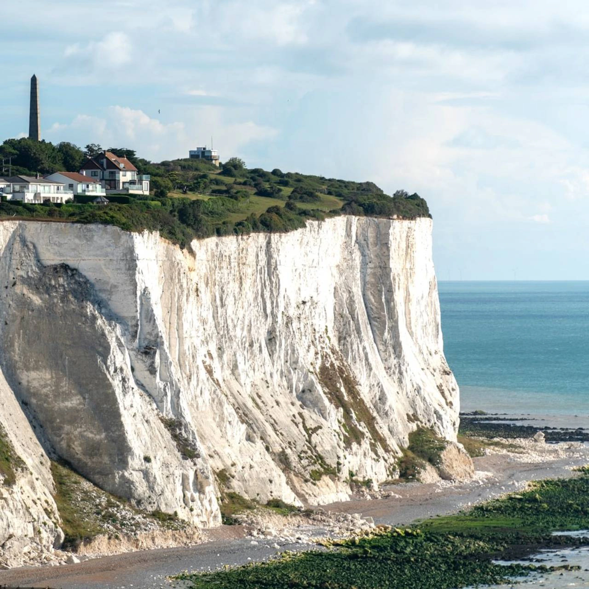 White cliffs along a shoreline with houses and a tall monument on top, meeting the calm blue sea under a partly cloudy sky.