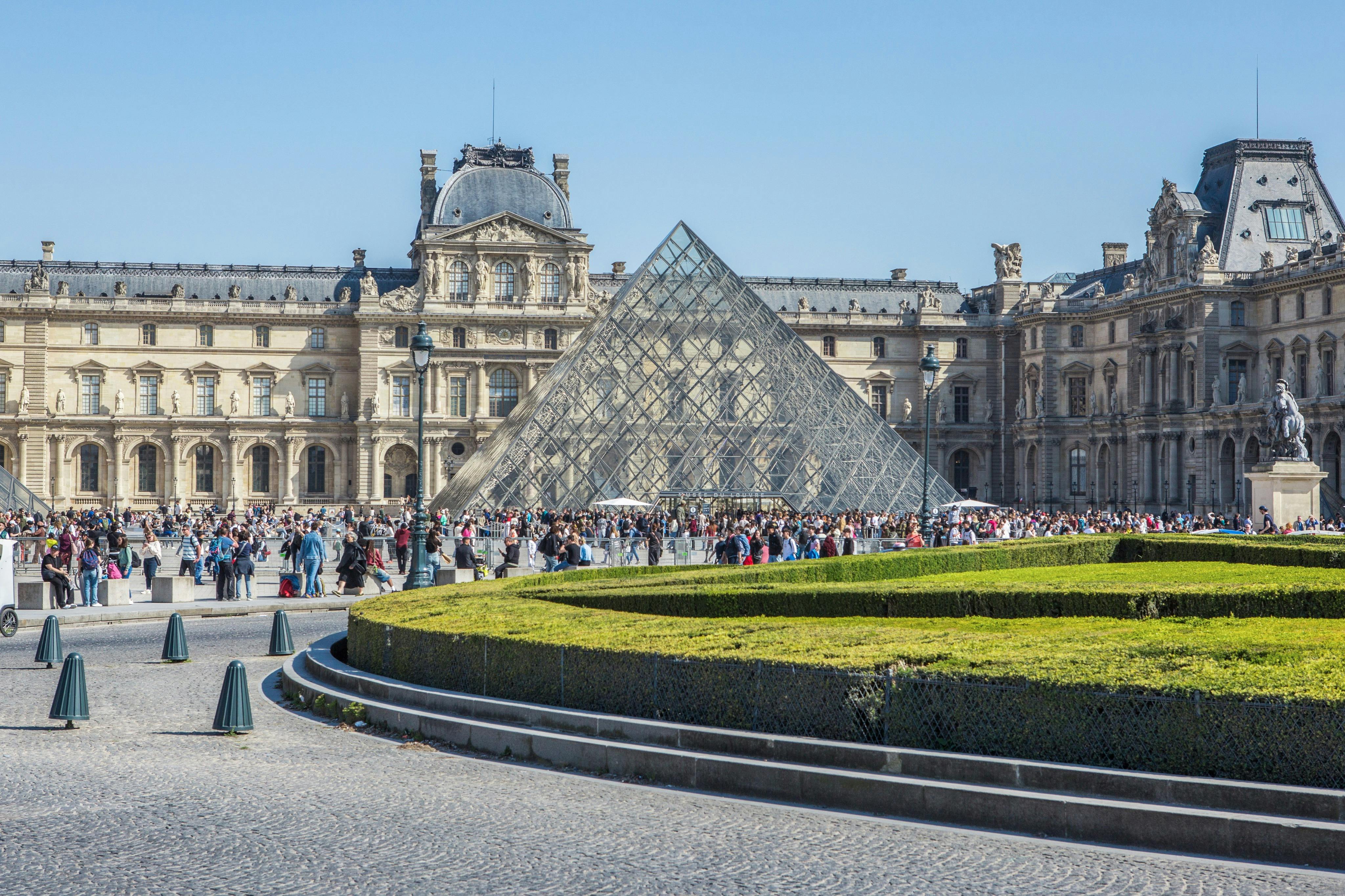 Skleněná pyramida s historickou budovou v pozadí, obklopená davy lidí a upravenou zahradou v Paříži.