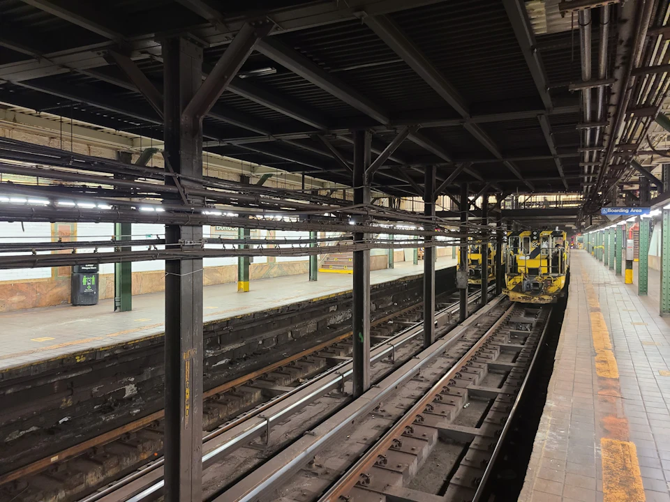An empty train station platform with tracks, overhead beams, and maintenance equipment visible in the distance.