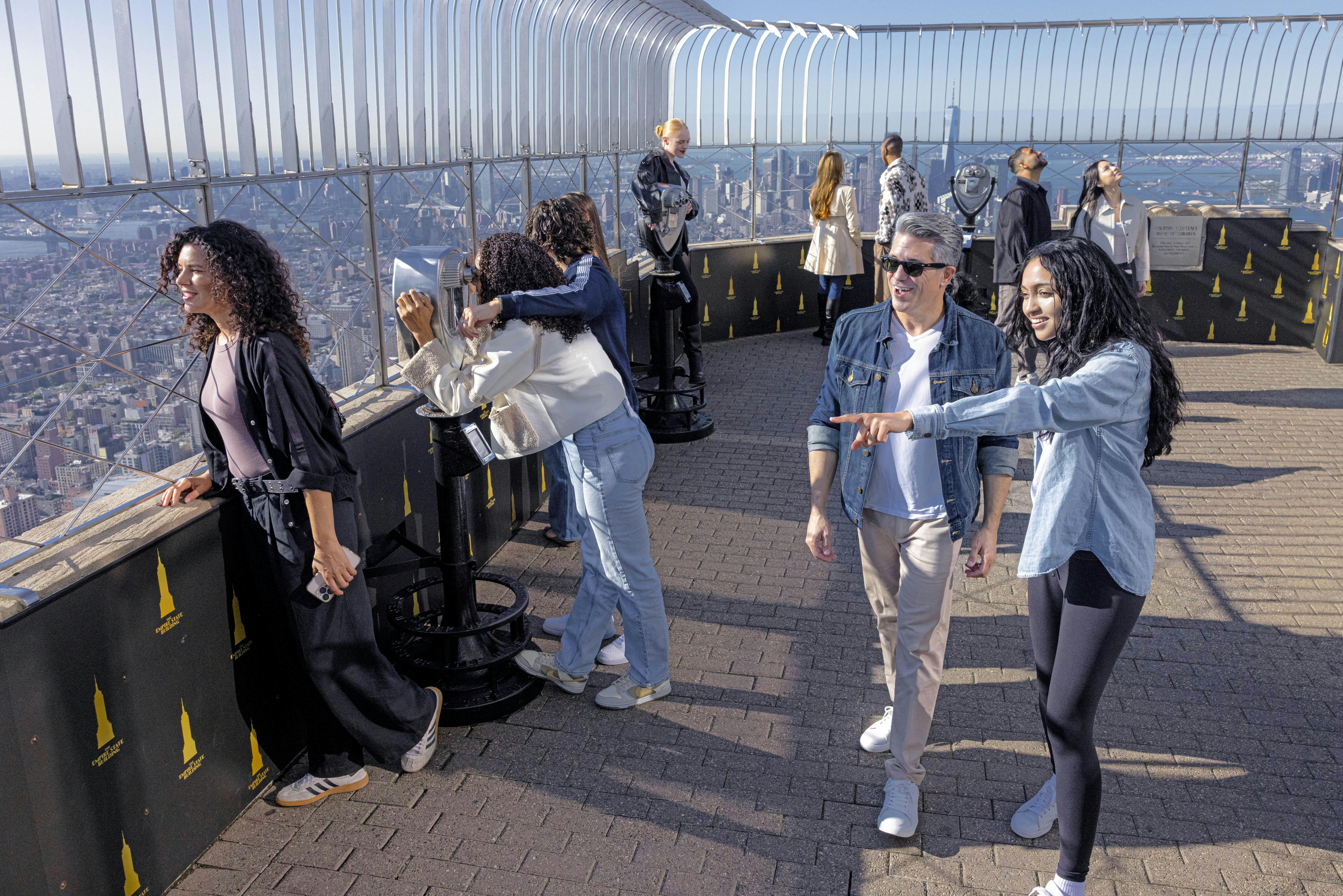 People enjoying the view from an observation deck with telescopes and safety railings on a sunny day.