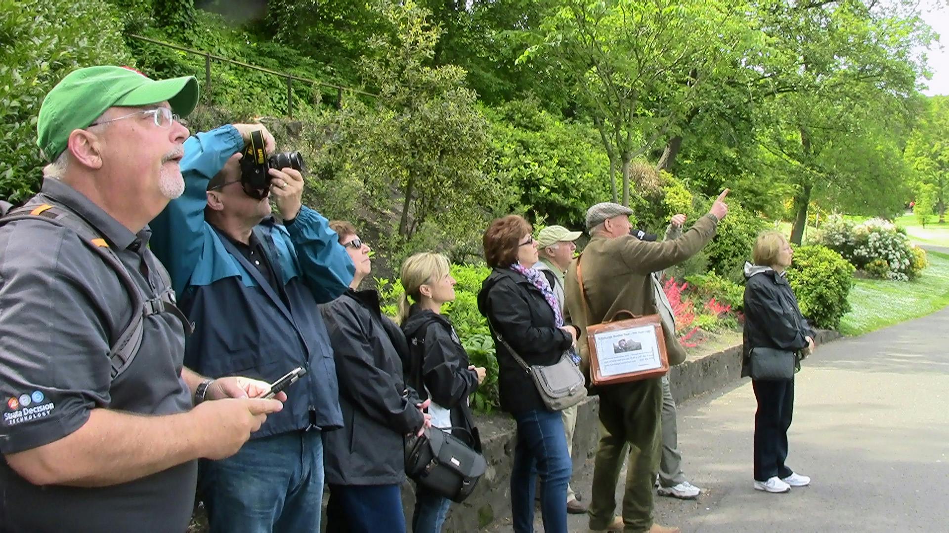 Un gruppo di persone all'aperto, alcune delle quali utilizzano macchine fotografiche per fotografare l'ambiente circostante, con alberi e arbusti verdeggianti sullo sfondo.