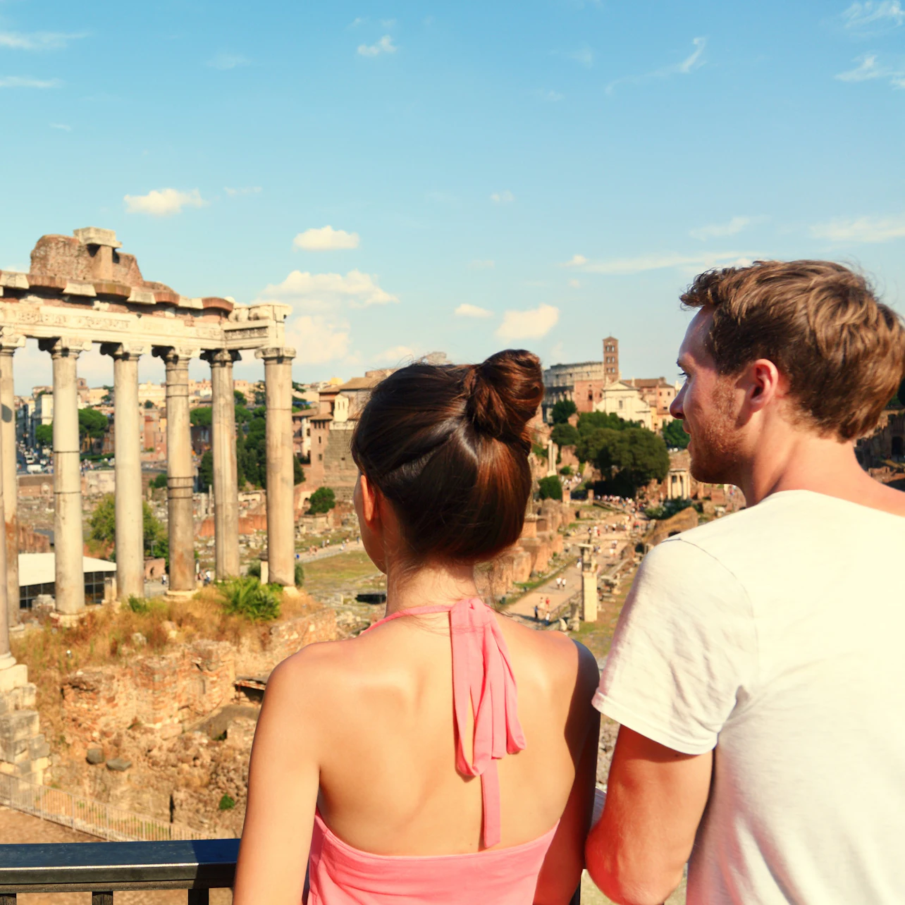 Two people facing away, overlooking ancient ruins with stone columns under a clear blue sky.