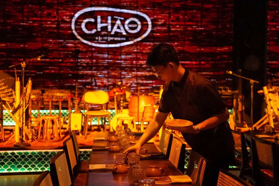 A person sets a table in a dimly lit restaurant with "CHÁO" illuminated on the brick wall in the background.