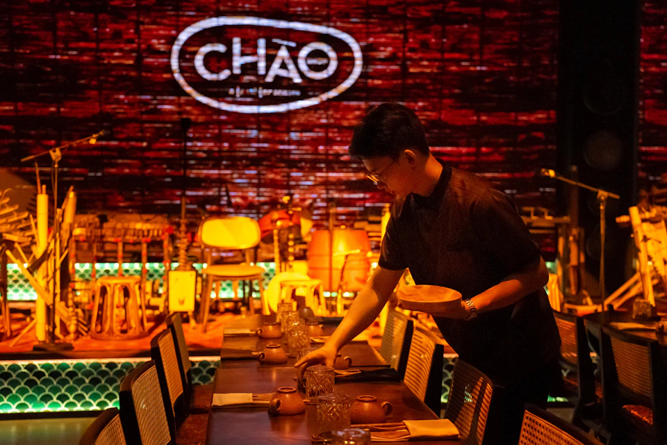 A person sets a table in a dimly lit restaurant with "CHÁO" illuminated on the brick wall in the background.