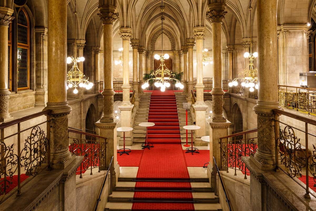 Μεγάλη σκάλα με κόκκινο χαλί, περίτεχνους πολυελαίους, μαρμάρινες κολώνες και στρογγυλά τραπέζια σε μια πολυτελή αίθουσα.