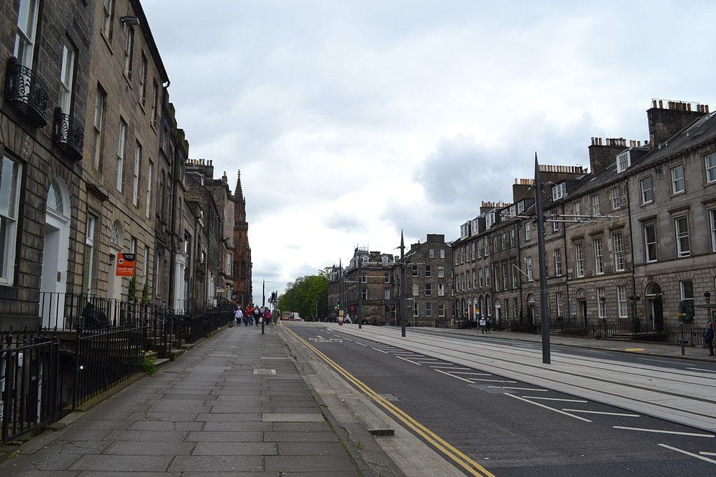 A street with stone buildings on both sides, tram tracks in the road, a few people walking on the sidewalk, and overcast skies.