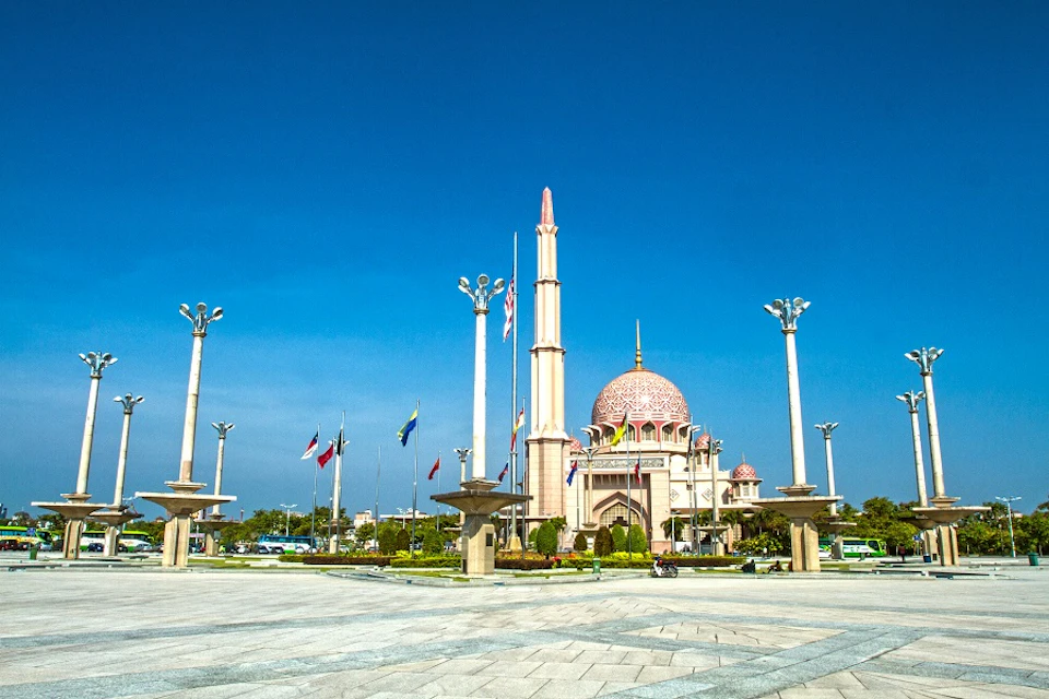 Ornate pink-domed mosque with tall minaret, surrounded by flags and lamp posts, under a clear blue sky.