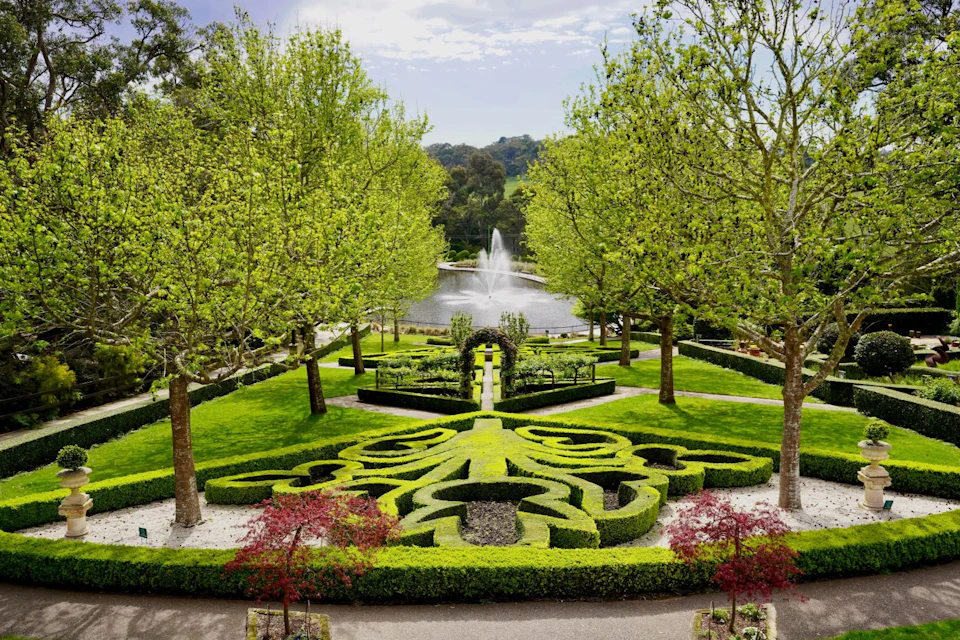A formal garden with intricate hedges, a fountain in the background, and tall trees surrounding the landscape.