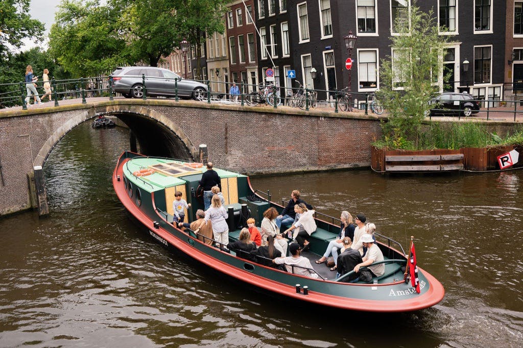 Pessoas sentadas num barco de canal em Amesterdão, passando por baixo de uma ponte com bicicletas estacionadas e edifícios em segundo plano.