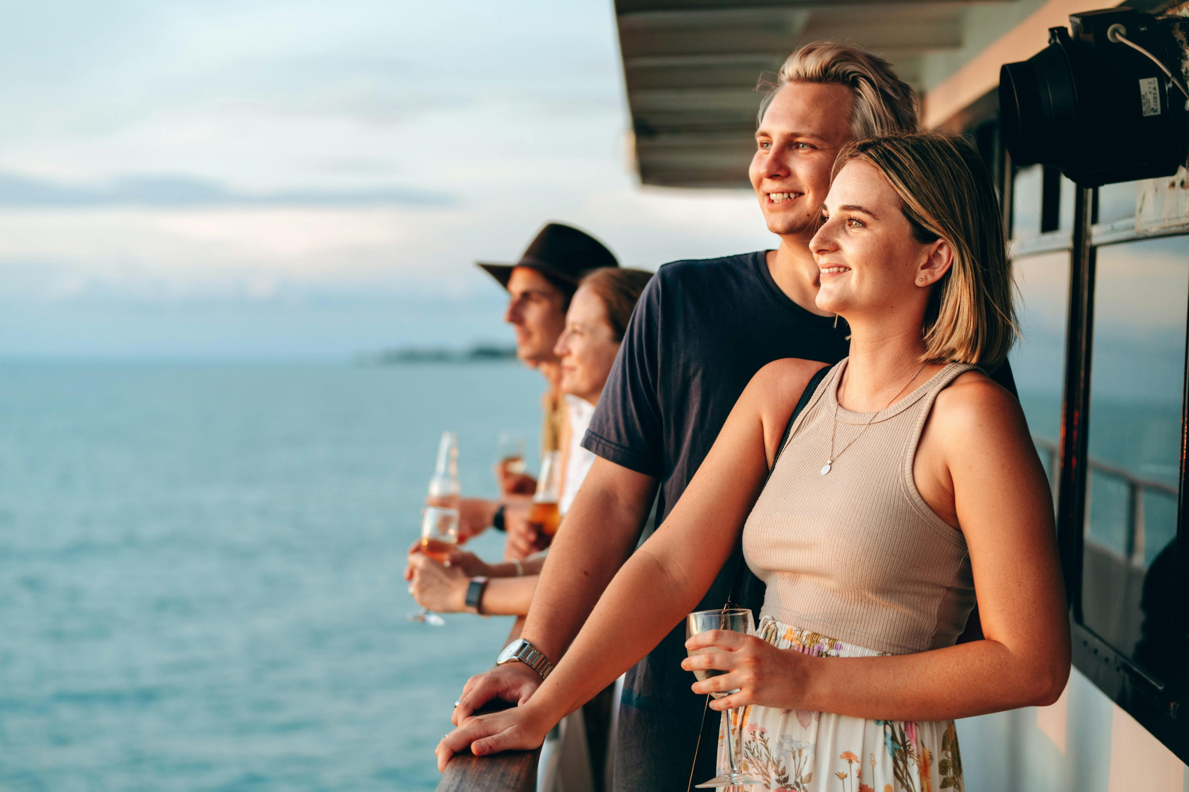 Four people standing on a deck, holding drinks, and looking out over a body of water during sunset.