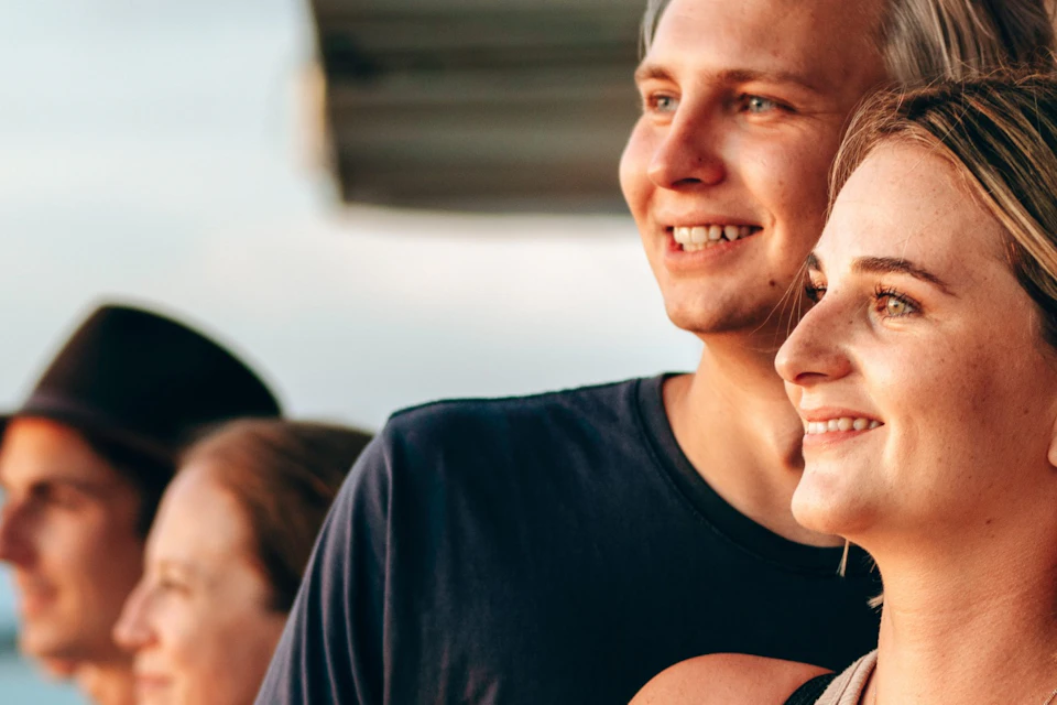Four people standing on a deck, holding drinks, and looking out over a body of water during sunset.