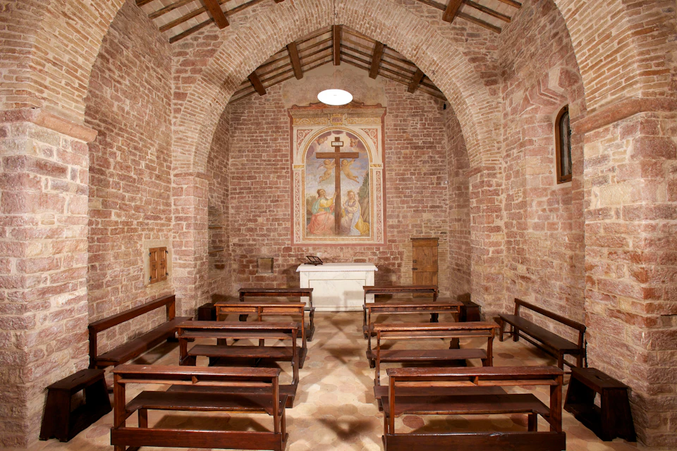 Small chapel with stone walls, wooden pews, and a painting of the crucifixion behind a white altar.