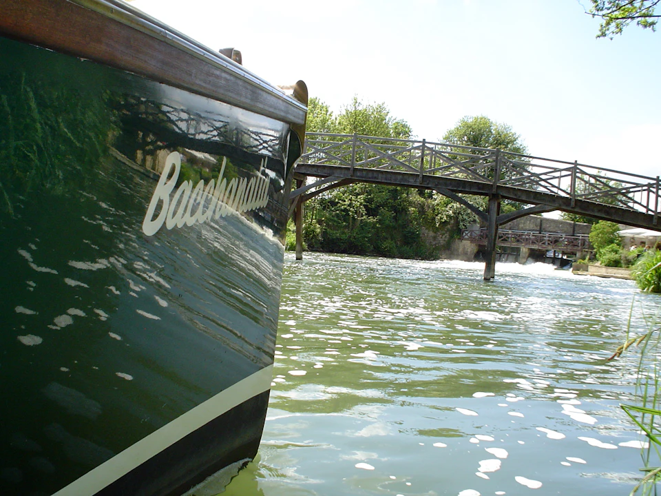 Boat named "Bacchagani" on a waterway with a wooden footbridge and greenery in the background.
