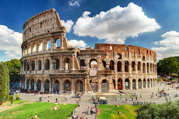 Colosseum i Rom under en klarblå himmel med spridda moln, omgiven av turister som går och fotograferar.