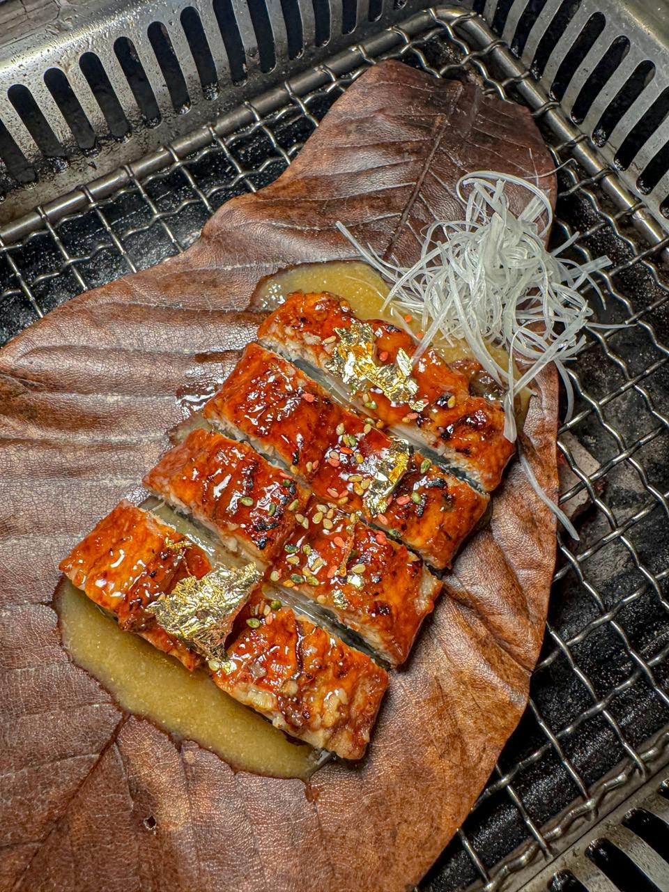 Grilled glazed eel slices with gold leaf garnishes on a leaf, served with shredded white garnish on a grill surface.