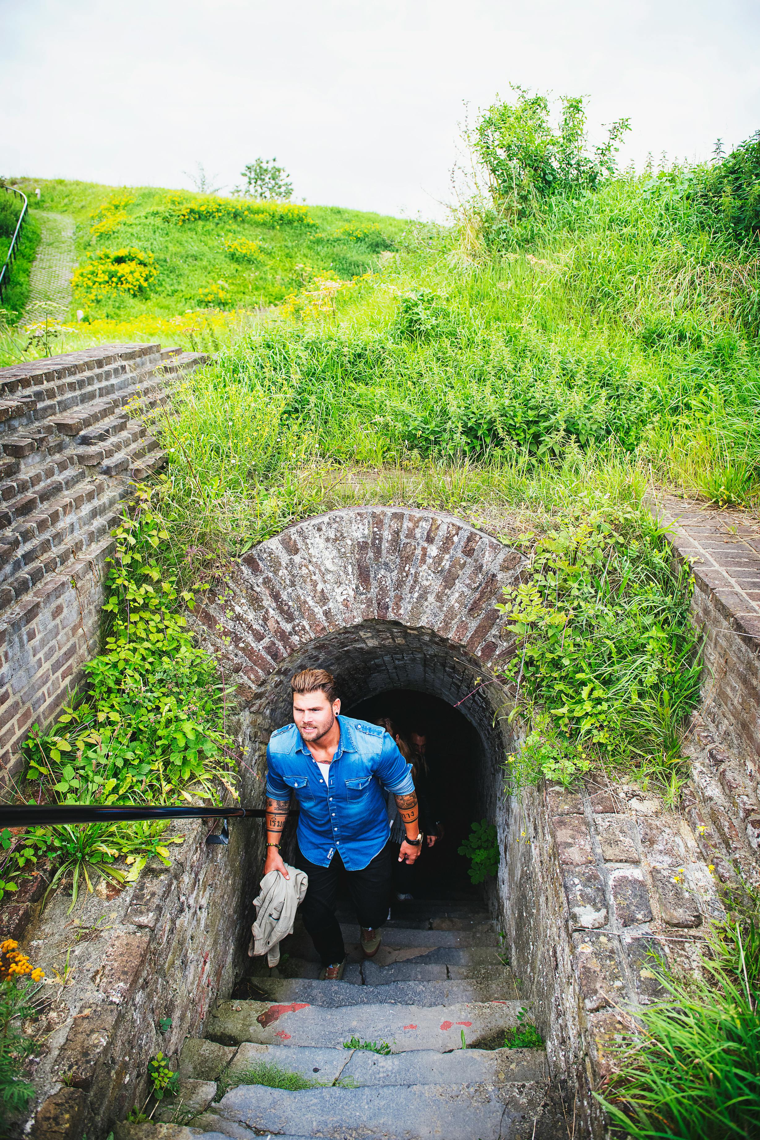 En man i blå skjorta kommer ut ur en stentunnel omgiven av grönska en solig dag.