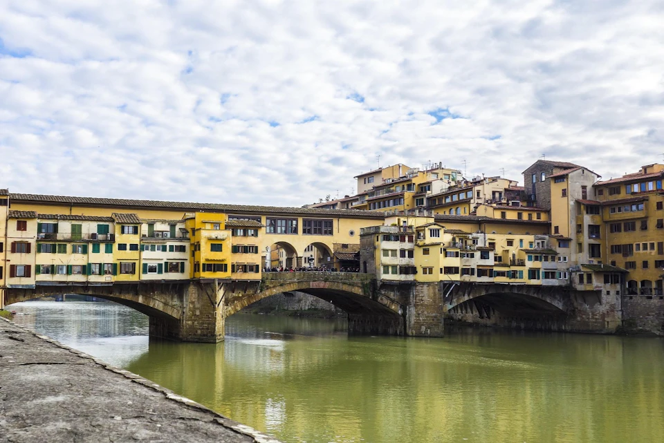A historic bridge with buildings on it spans a river under a cloudy sky.