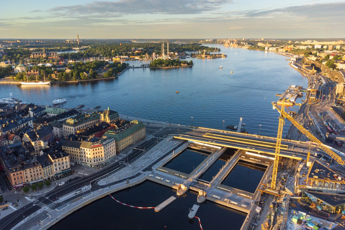 Aerial view of a waterfront city area with buildings, roads, canals, and a large body of water under a partly cloudy sky.