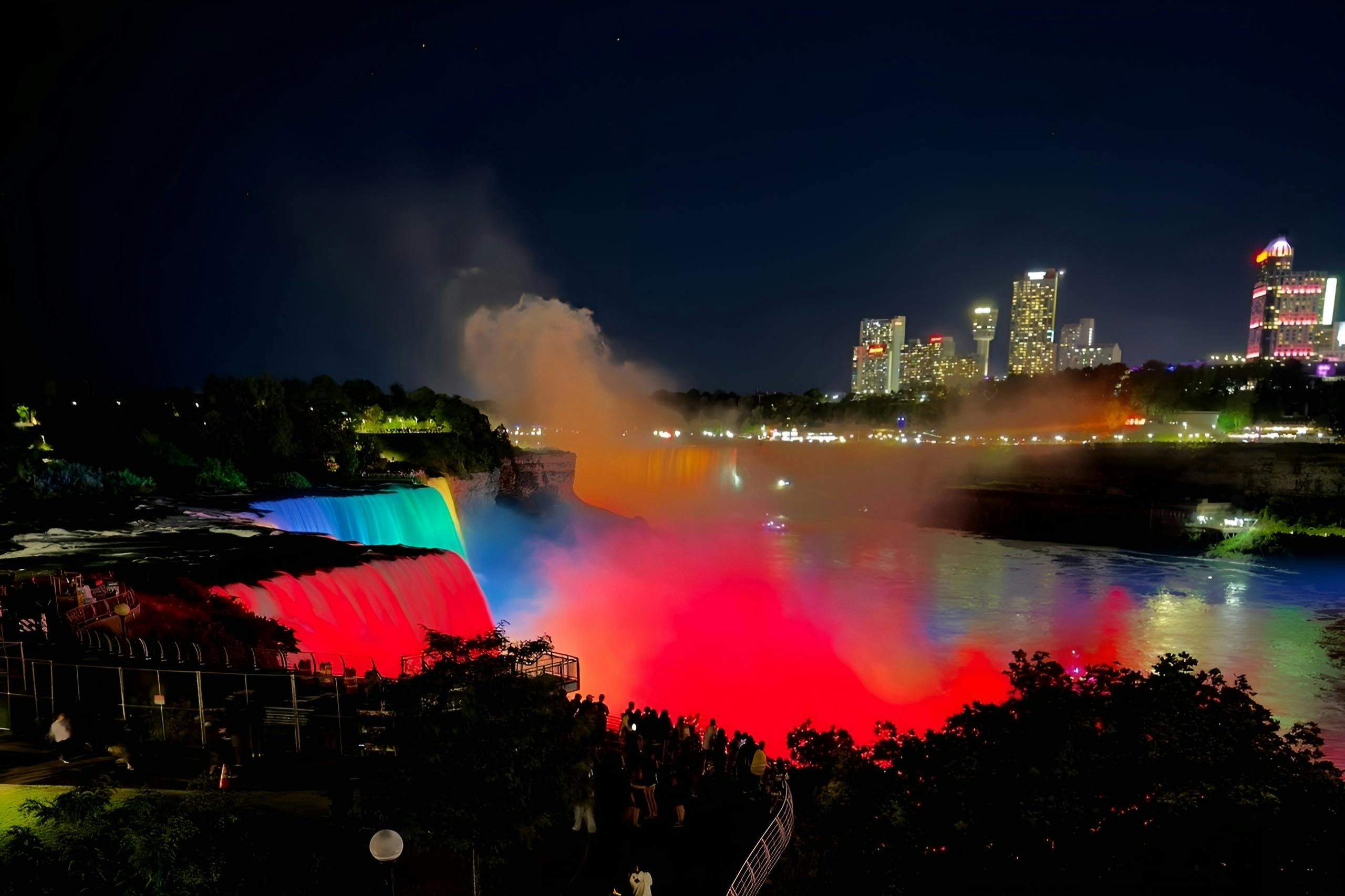 夜景中的尼亚加拉大瀑布灯光五彩缤纷，前方有城市建筑和一群人。