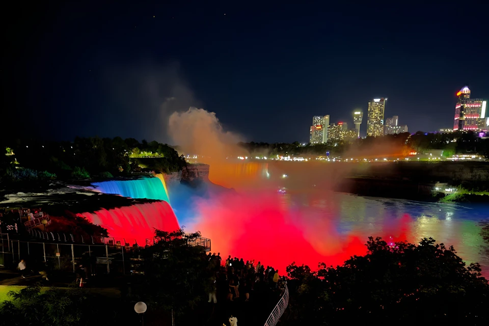 A night view of Niagara Falls illuminated with colorful lights, with city buildings and a crowd of people in the foreground.