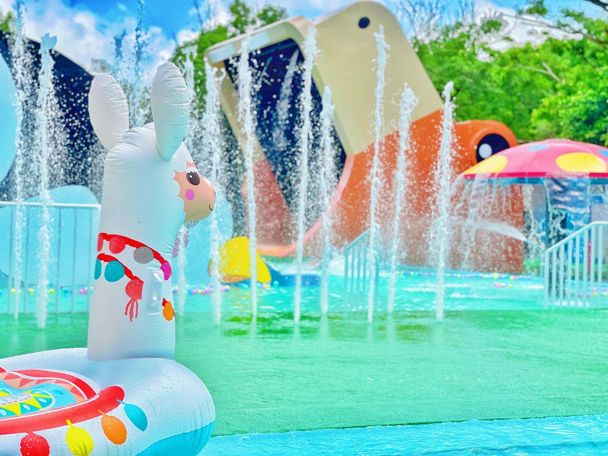 A colorful inflatable llama float in a water park with fountains, slides, and greenery in the background.