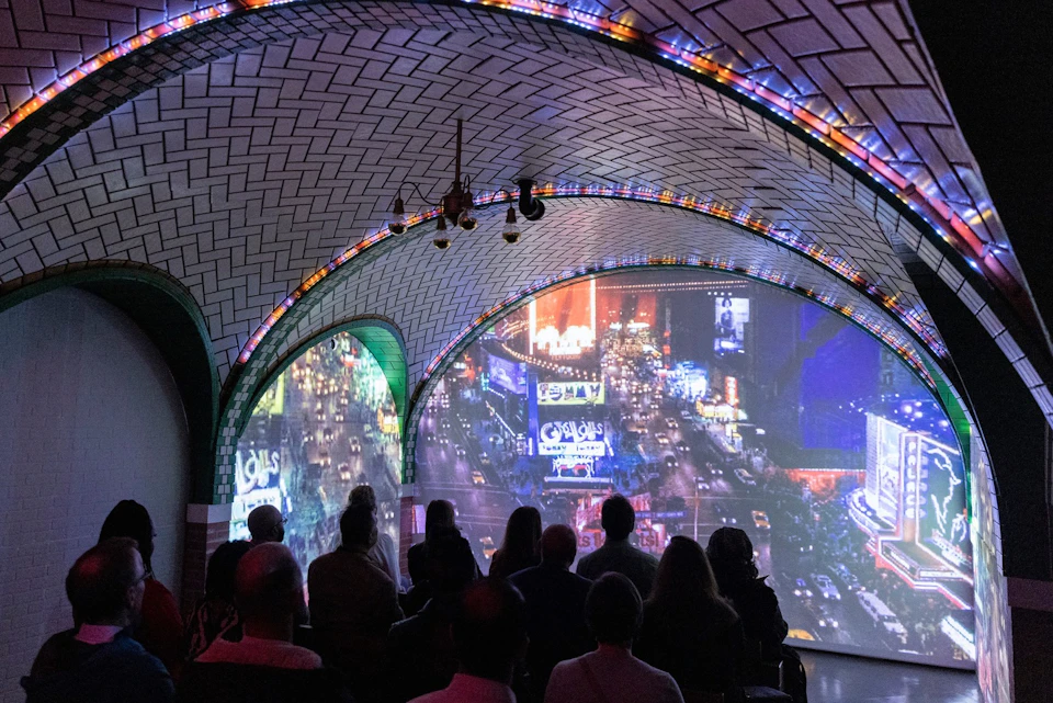 People seated in a dimly lit room with arched ceilings, watching a large projection of a city street at night.