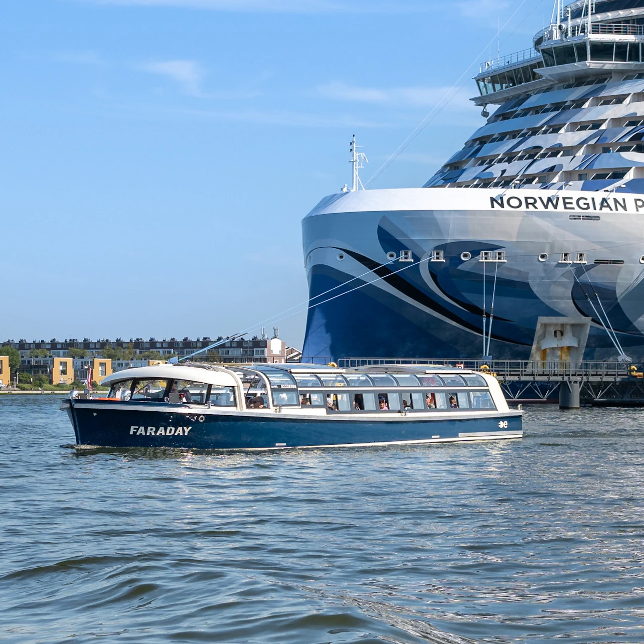 A small blue passenger boat named Faraday on water near a large cruise ship with 'Norwegian' visible on its hull.