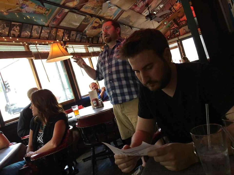 Four people in a dimly-lit restaurant, one man standing and gesturing, another reading a paper, two seated and conversing.