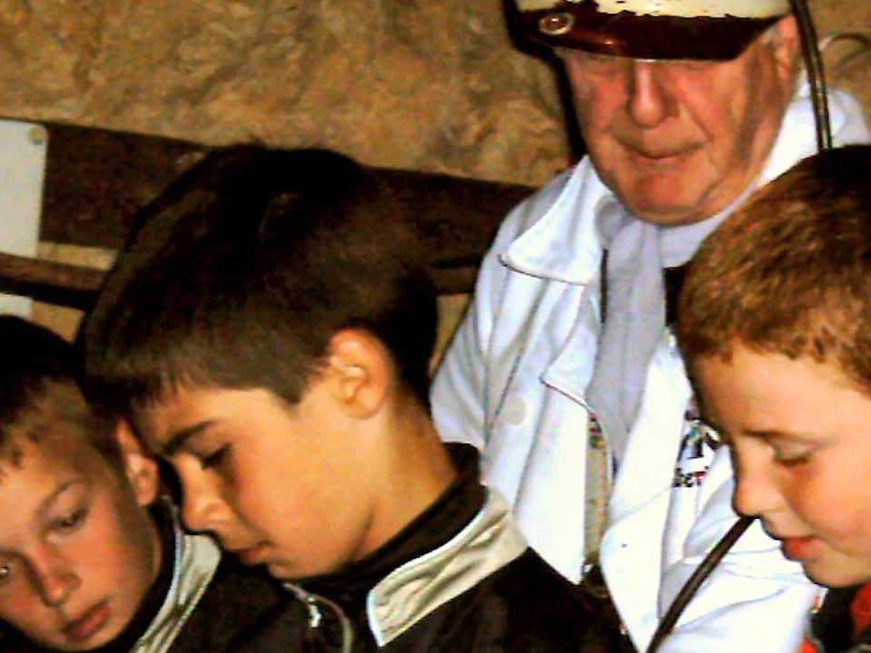 Four boys, supervised by an adult in a white coat, explore a mechanical device with pipes and valves in a dimly lit room.